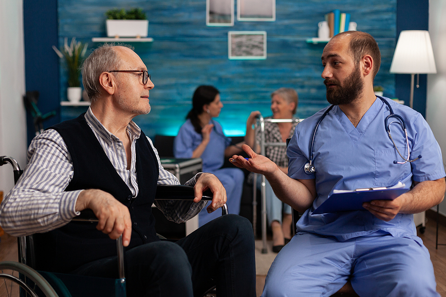 Male medical assistant sitting next to elderly patient in a waiting room explaining the papers on his clipboard.
