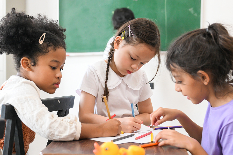 three young girls sitting at a table in a classroom coloring with colored pencils