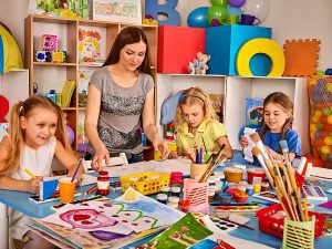 Teacher standing at a table, while leading an art activity to a group of kids. 