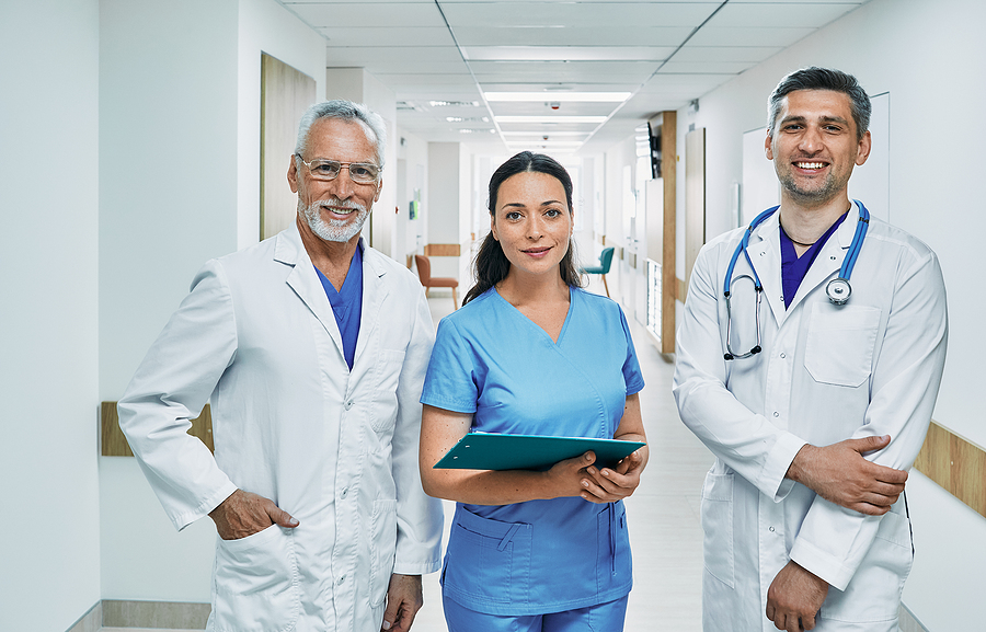 Medical team including a doctor, nurse, and medical assistant standing in a hospital corridor. Medical assistant is wearing blue scrubs and holding a blue clipboard.