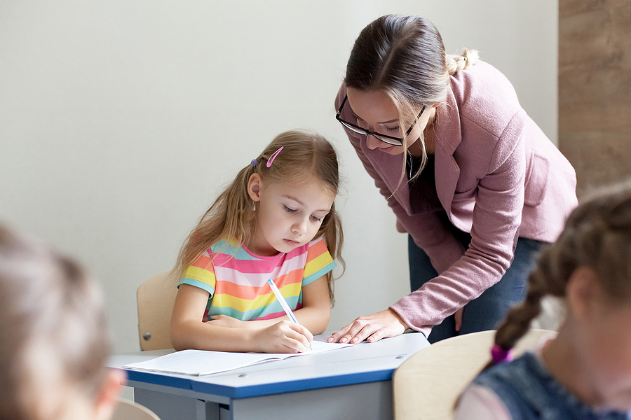Female elementary school teacher standing next to a girl's desk helping her with her work