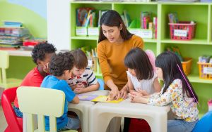 Female teacher sitting at a table, helping a group of young students. 