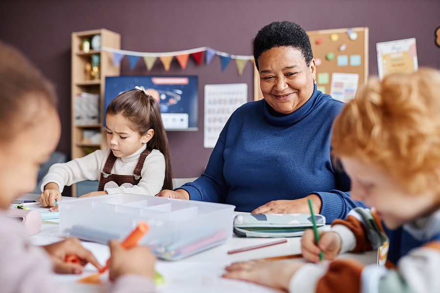 Female teacher with short hair sitting with her preschool students while they draw and color.