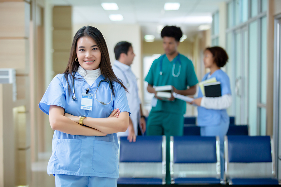 Woman in scrubs smiling standing in medical wait room with other medical staff having a conversation in the background