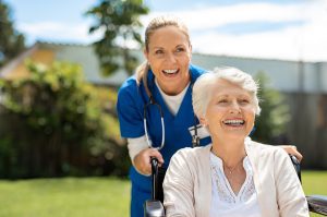 Happy female medical assistant and elderly woman sitting in wheelchair smiling in an outdoor setting. 