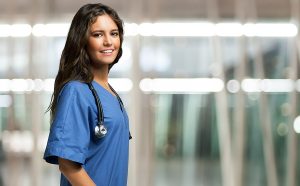 Smiling female medical assistant wearing blue scrubs with a blurred out background behind her. 