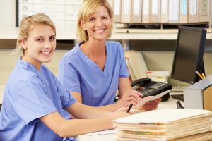Two female medical workers wearing blue scrubs and sitting at a desk with medical files. 