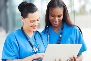 Two female nurses wearing blue scrubs and looking at a laptop smiling in a medical setting. 