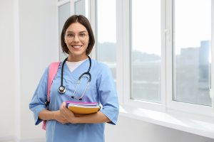 Female nursing student standing in front of window holding a stack of books. 
