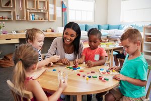 Female teacher sitting at a wood table with a group of young students leading a lesson. 