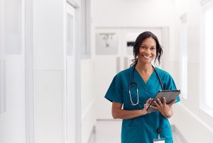 Female nurse wearing green scrubs, holding a clipboard in the hallway of a medical facility. 