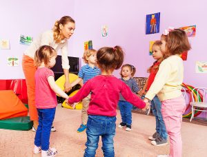 Female teacher holding hands with a group of students standing in a circle in a bright, colorful classroom setting. 