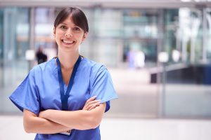 Female nurse wearing blue scrubs and standing outside a medical facility. 