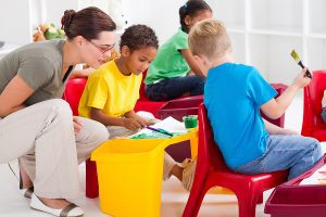 Female preschool teacher helping kids paint pictures. 