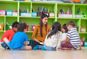 Female teacher sitting on a classroom floor reading to a group of young students. 
