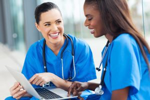 Two female nurses wearing blue scrubs smiling and talking in front of an open laptop. 
