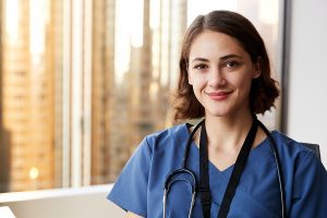 Female nurse wearing blue scrubs, standing in front of a hospital window smiling. 