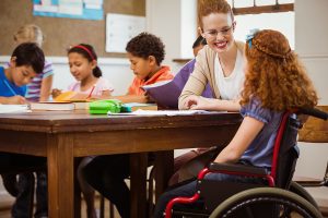 Female teacher helping a group of students seated at their desk and one girl in a wheelchair. 