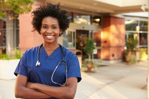 Smiling female nurse wearing blue scrubs, smiling in front of a hospital. 