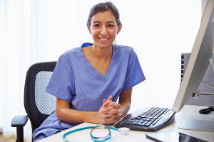 Female nurse wearing blue scrubs, sitting in front of a computer, smiling. 