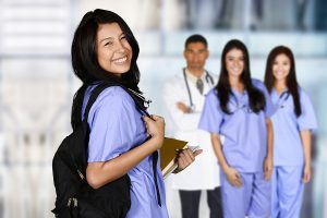 Smiling female nurse wearing a black backpack and scrubs with a doctor and fellow nursing students behind her. 