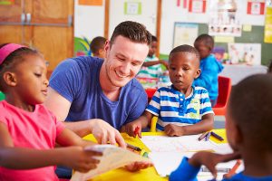 Male teacher working with a group of young students at their desks. 