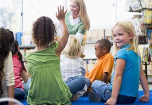Female teacher standing at the front of a classroom teaching a group of young students. 