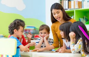 Female preschool teacher standing in classroom, reading to a group of young children. 