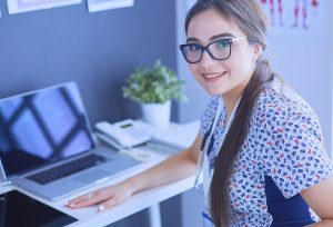 Female healthcare professional wearing scrubs and sitting in front of a laptop in a medical office.  
