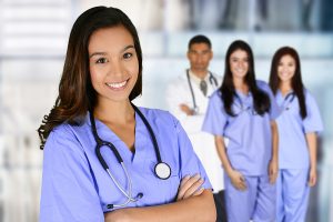 Nurse wearing blue scrubs smiling and standing in front of a group of medical professionals. 