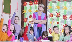 Female preschool teacher standing in her classroom with paper flowers on the windows and a group of young children sitting at the table. 