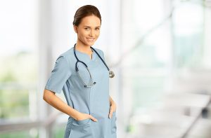 Female nurse standing in front of a window with her hands in her pockets, wearing light blue scrubs. 