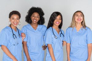 Group of four female nurses, wearing blue scrubs and smiling, standing next to each other. 