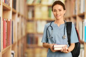 Female nursing student wearing light blue scrubs, a backpack and stethoscope, standing in a library with books in her hands. 