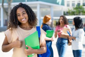 Smiling female adult student, holding a folder and standing in front of other students giving a thumbs up. 