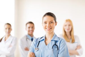 Female nurse wearing blue scrubs and standing in front of other medical professionals.