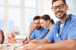 Male nursing student wearing blue scrubs, sitting in a classroom with other nursing students. 