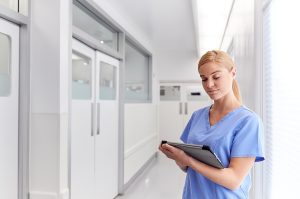 Female nurse wearing scrubs and standing in a hospital corridor using a tablet. 