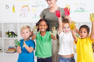 Female preschool teacher standing in her classroom with four preschool students who have paint on their hands. 