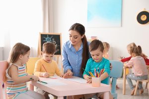 Female preschool teacher sitting at a table with her young students in a classroom.