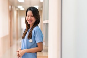 Female nurse wearing light blue scrubs and smiling in the hallway of a medical facility 