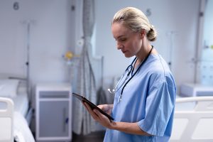 Female nurse wearing blue scrubs and staring down at a tablet in a hospital room. 