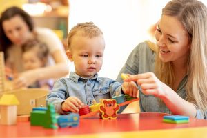 Childcare teacher sitting at a table with a toddler, helping him with an activity.