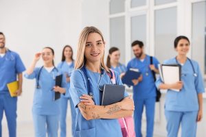 Female nursing student wearing blue scrubs and holding books in her arms, standing in front of other nursing students.