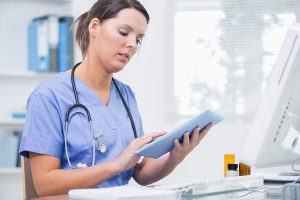 Female nurse wearing light blue scrubs and typing on a digital tablet. 