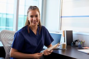 Female nurse wearing dark blue scrubs, sitting at a desk, smiling.