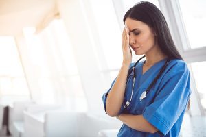 Female nurse wearing blue scrubs and holding her hand up to her head, looking stressed in a medical facility.