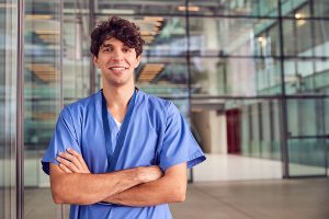 Male nursing wearing blue scrubs and standing outside a medical facility.