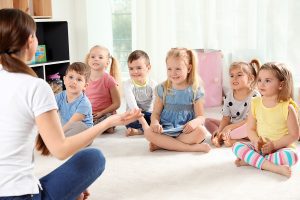 Preschool teacher sitting on the floor, leading an activity to a group of students.