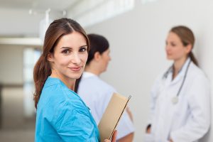 Female nurse holding a patient chart and smiling with a doctor and patient in the background.